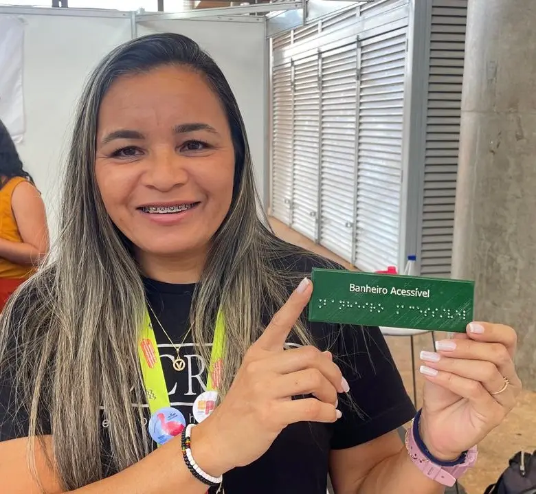 Uma mulher loira participante de feira científica segura uma placa verde com escrita em braille que indica “Banheiro Acessível”. Ela sorri e aponta para o item, destacando o foco em acessibilidade. No fundo, colegas interagem no estande do IFCE.