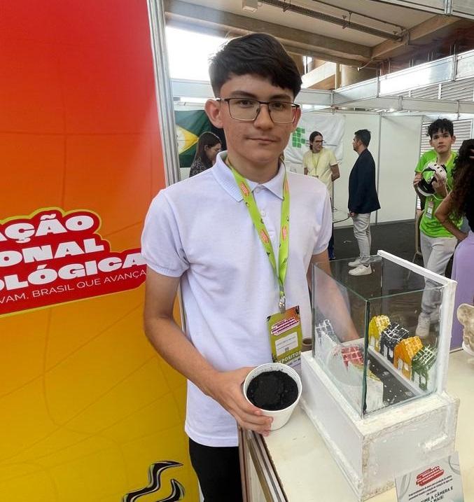 Um estudante magro e de óculos apresenta um protótipo de asfalto ecológico em uma maquete transparente, exibindo a simulação da rua de uma cidade. Ele segura um recipiente com amostra do material. O estande possui banner laranja com o tema da Educação Profissional e Tecnológica.