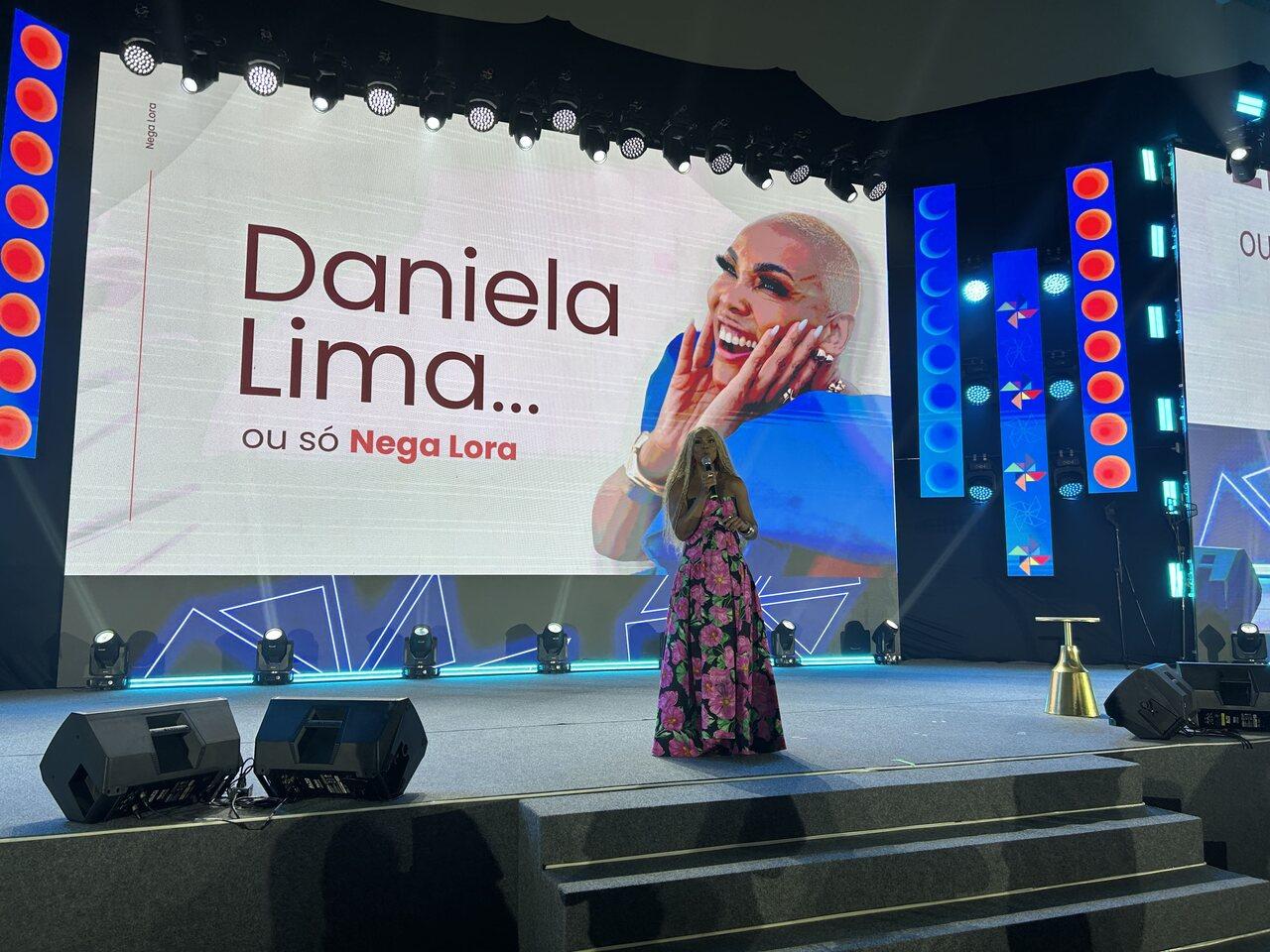 Daniela Lima, conhecida como Nega Lora, durante palestra em evento de empreendedorismo, diante de telão com seu nome e imagem ao fundo.