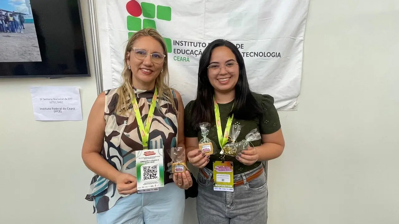 Duas mulheres de óculos exibem produtos naturais em um estande do Instituto Federal do Ceará (IFCE). Elas seguram pequenos pacotes de biscoitos e frascos de óleos essenciais, com crachás de expositoras. Atrás delas, há o logotipo do IFCE em um banner branco.