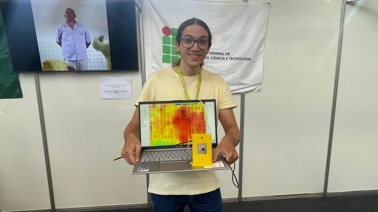 Um jovem demonstra um sistema de detecção de quedas com um notebook e um sensor amarelo. A tela exibe um mapa térmico colorido, indicando áreas de movimento. O projeto é apresentado no estande do IFCE, identificado por seu banner institucional.