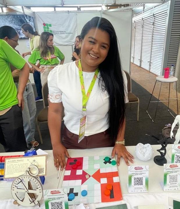Uma estudante apresenta um jogo de tabuleiro colorido voltado ao ensino da matemática. A mesa exibe peças educativas, modelos anatômicos e QR Codes para informações adicionais. Ela usa crachá de exposição e sorri diante do estande.