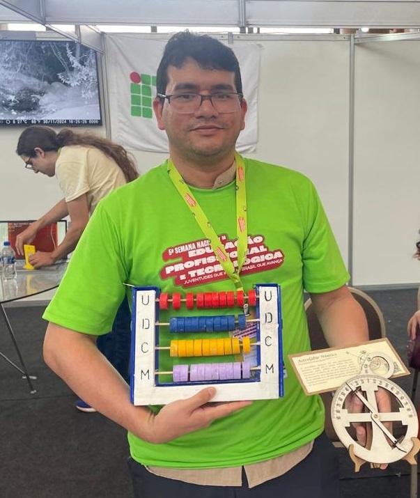 Um expositor de óculos e blusa verde segura instrumentos pedagógicos criados em laboratório maker, como um ábaco e um modelo de engrenagem matemática. Ele veste camiseta verde do evento e crachá de expositor. O fundo mostra colegas e o banner do IFCE.