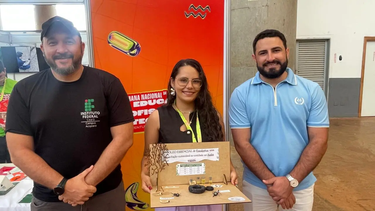 Três participantes de feira científica, incluindo uma mulher ao centro, apresentam um projeto sobre óleos essenciais de eucalipto usados no combate à dengue. O grupo do IFCE segura uma maquete explicativa com amostras e informações sobre o produto. O estande tem fundo laranja vibrante.