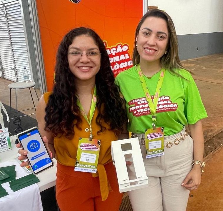 Duas expositoras do IFCE apresentam um projeto que utiliza inteligência artificial para analisar alimentos. Uma delas segura um celular com o aplicativo aberto, enquanto a outra exibe um protótipo branco que integra o sistema. Elas usam crachás de exposição e estão diante de um painel laranja da Semana Nacional de Ciência e Tecnologia.