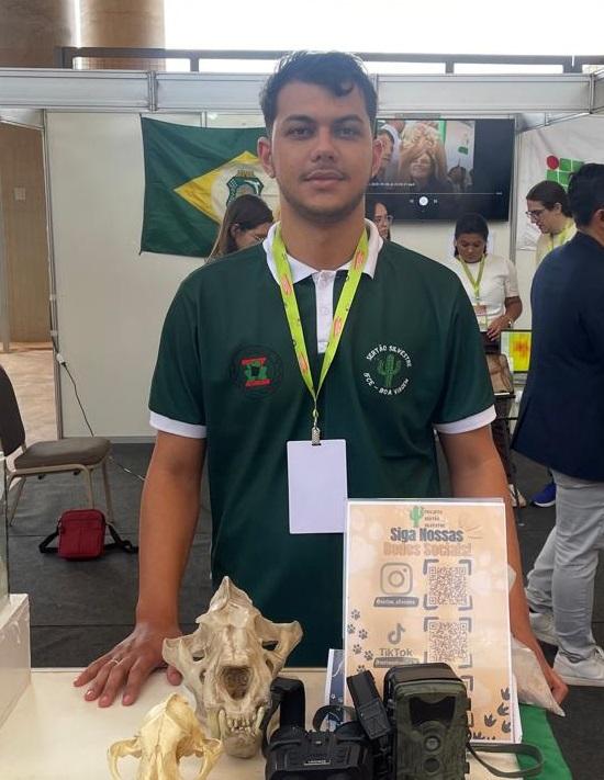 Aluno do IFCE com blusa verde e crachá posa em frente a bancada com crânios de animais, binóculos, câmeras e um informativo do projeto Sertão Silvestre, que monitora fauna no sertão do Ceará.