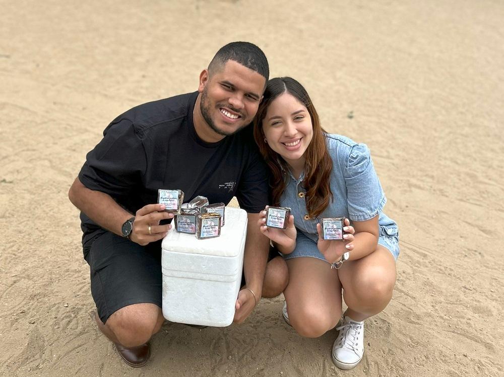 Na imagem, uma foto de arquivo pessoal mostra Ezequiel e Larissa, um casal jovem, agachados em uma área de terra ou areia. Ezequiel, à esquerda, usa uma camiseta preta e shorts pretos. Larissa, à direita, usa um macacão jeans de manga curta. Ambos sorriem para a câmera. Eles seguram pequenos objetos embrulhados, provavelmente sabonetes artesanais ou produtos semelhantes, e uma pequena caixa térmica branca está entre eles, sobre a qual há mais desses pequenos embrulhos.