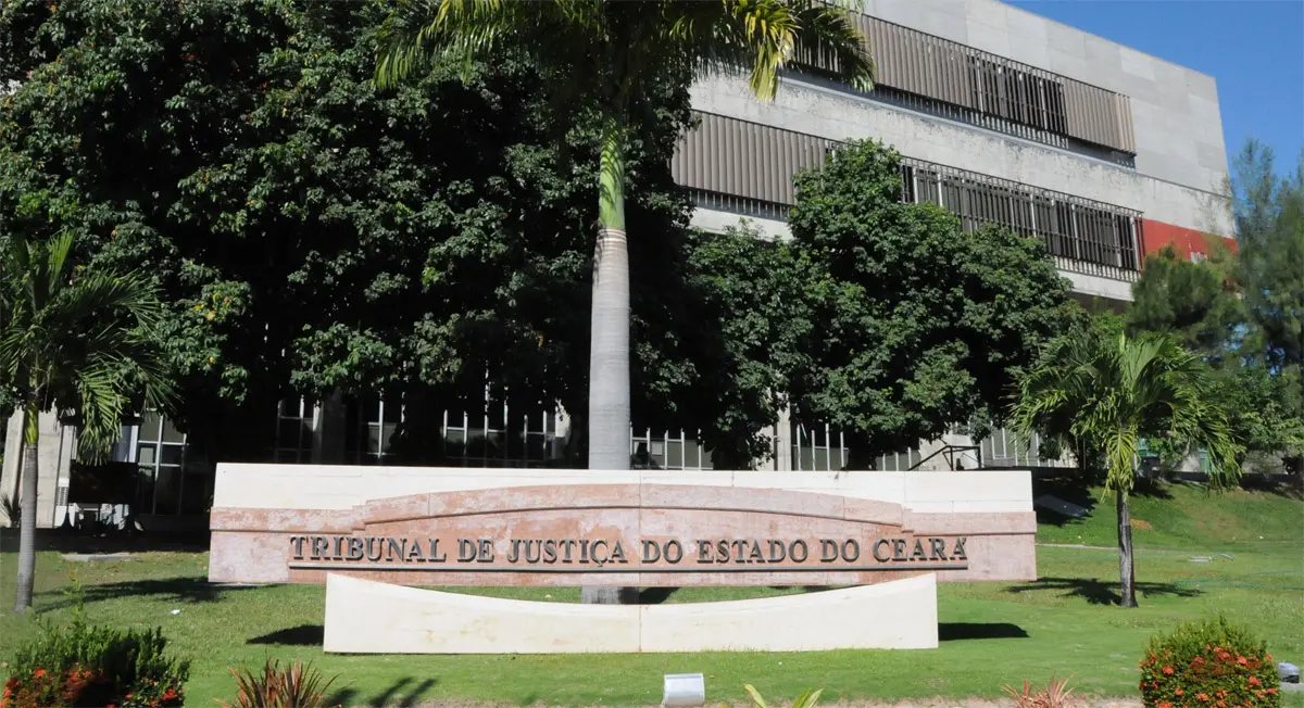 Fachada do Tribunal de Justiça do Estado do Ceará rodeada por árvores e jardim, com uma placa de mármore na frente. Edifício moderno com design contemporâneo.