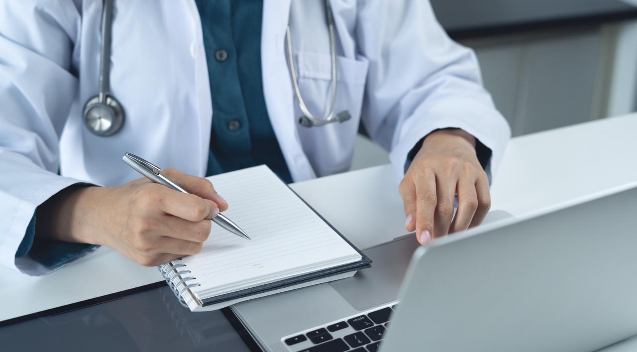 Médico trabalhando com laptop e anotando informações em uma clínica ou consultório, representando cuidado com saúde e atendimento médico.