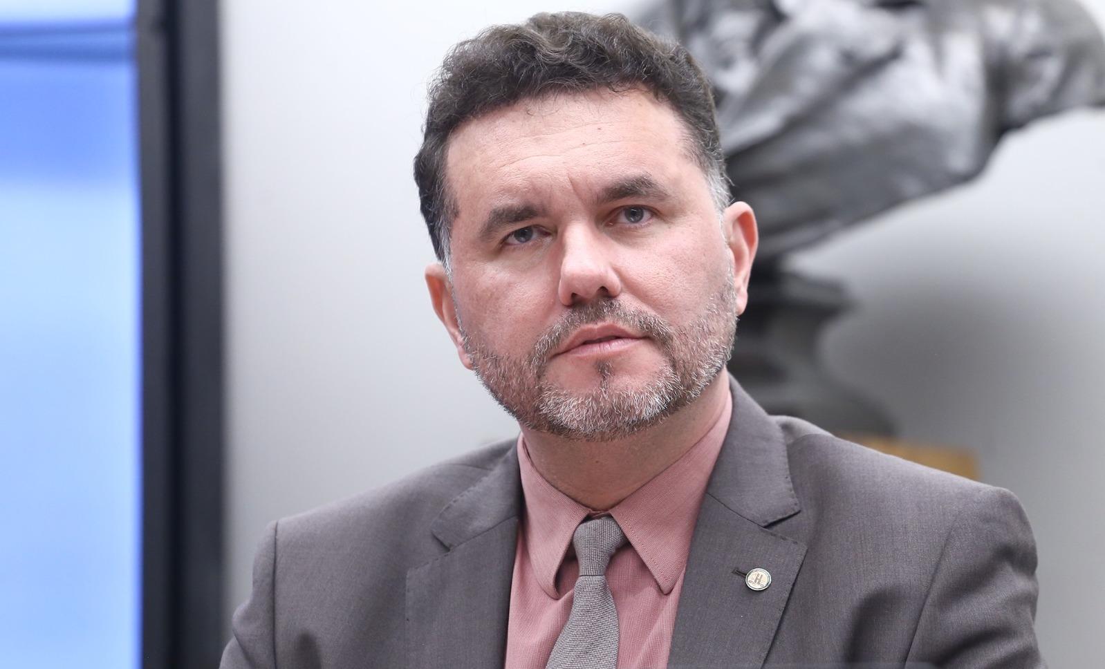 Moses Rodrigues é um homem de meia idade com barba e cabelo grisalhos. Na foto, ele usa terno cinza com camisa social rosa e gravata cinza.