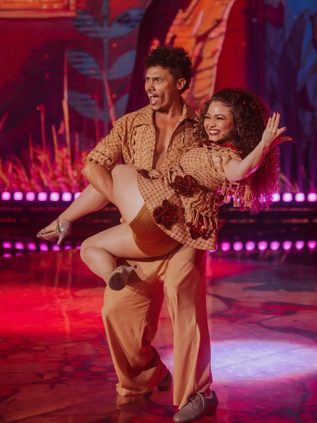 Na imagem, uma performance de dança. Um homem, com cabelo cacheado escuro, pele bronzeada e vestindo uma camisa marrom-claro de manga curta e calças combinando, segura uma mulher em seus braços. A camisa dele está aberta no peito. A mulher tem cabelo cacheado escuro, um sorriso largo e veste um body ou roupa de banho marrom-dourado com detalhes em franjas e apliques vermelhos na parte superior. Ambos estão olhando para a frente ou ligeiramente para a esquerda do quadro com expressões intensas. O homem está levantando a mulher de forma dramática, e ela tem um braço levantado. Eles estão em um palco com luzes de palco em tons de rosa, roxo e vermelho e um fundo que parece ser uma projeção de folhagens estilizadas. O nome do arquivo sugere que o homem é Silvero Pereira e a foto é da 