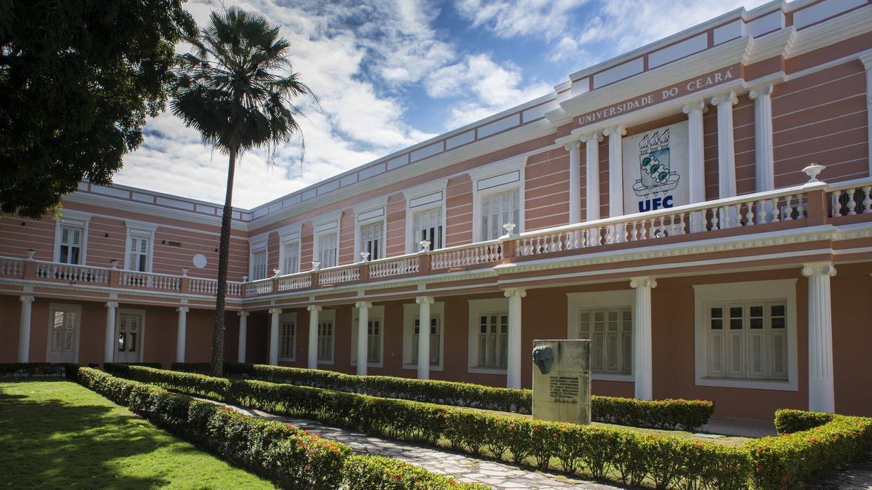 Fachada da Universidade Federal do Ceará. É um prédio antigo que mescla as cores branco e salmão e localizado num espaço com jardim.
