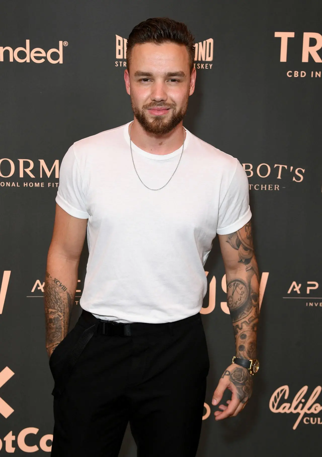 foto de Liam Payne, cantor que faleceu em outubro de 2024. ele veste camiseta branca e sorri diante de painel escuro com nomes de marcas.