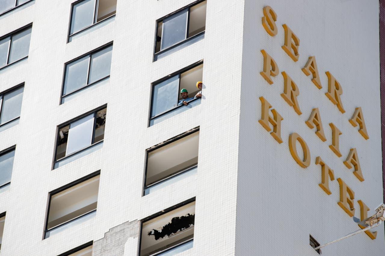 Fachada do Seara Praia Hotel vista de baixo, com trabalhadores usando capacetes apoiados em uma janela aberta e letras amarelas do nome do hotel na parede lateral.