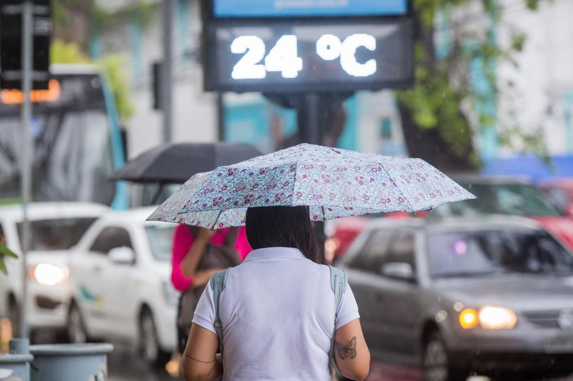 Uma mulher caminha sob a chuva com um guarda-chuva estampado, enquanto o termômetro de rua marca 24 °C. Ao fundo, carros e pedestres se movimentam entre o trânsito intenso do centro de Fortaleza. A cena retrata um dia nublado e de temperatura amena na capital cearense.