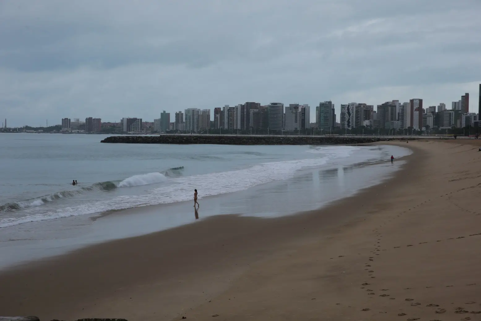 Vista ampla da orla de Fortaleza mostra a praia quase deserta em um dia cinzento. Algumas pessoas caminham à beira-mar e poucas se arriscam nas ondas suaves. Ao fundo, o horizonte urbano exibe a sequência de prédios da Avenida Beira-Mar sob o céu encoberto.