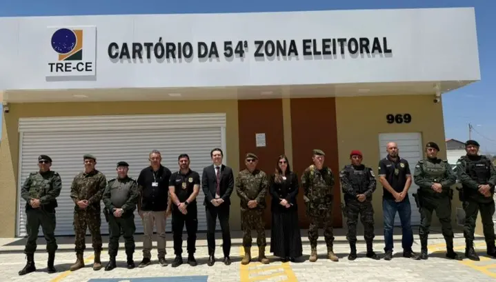 Imagem mostra um grupo de 13 pessoas posando em frente ao prédio do Cartório da 54ª Zona Eleitoral, do TRE-CE. O grupo é composto por homens e mulheres, a maioria vestida com uniformes militares ou policiais, e alguns com trajes formais. O prédio tem fachada clara, com o número 969 visível à direita e o logotipo do Tribunal Regional Eleitoral do Ceará acima da entrada. O céu está ensolarado.