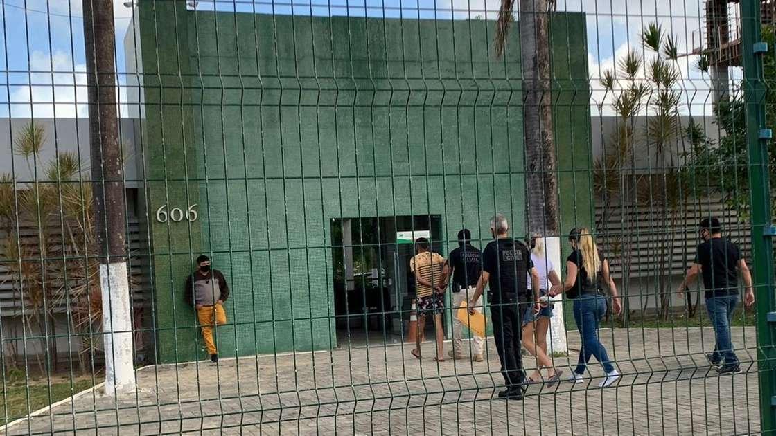 Grupo de pessoas entrando em uma unidade prisional ou penitenciária verde, vista através de uma cerca de grade metálica, indicando um ambiente de segurança.