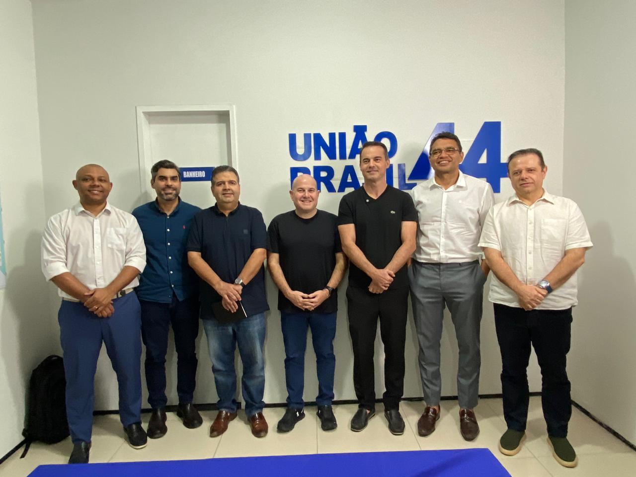 Sete políticos (Soldado Noélio, Queiroz Filho, Felipe Mota, Roberto Cláudio, Capitão Wagner, Sargento Reginauro e Cláudio Pinho) posam lado a lado em frente a uma parede branca com o logotipo do partido União Brasil e o número 44 em azul. Todos estão sorrindo e usam roupas sociais em tons neutros, variando entre camisas brancas, azuis e pretas. A foto parece ter sido feita em um ambiente interno de escritório, com piso claro e uma porta ao fundo com a placa “banheiro”.
