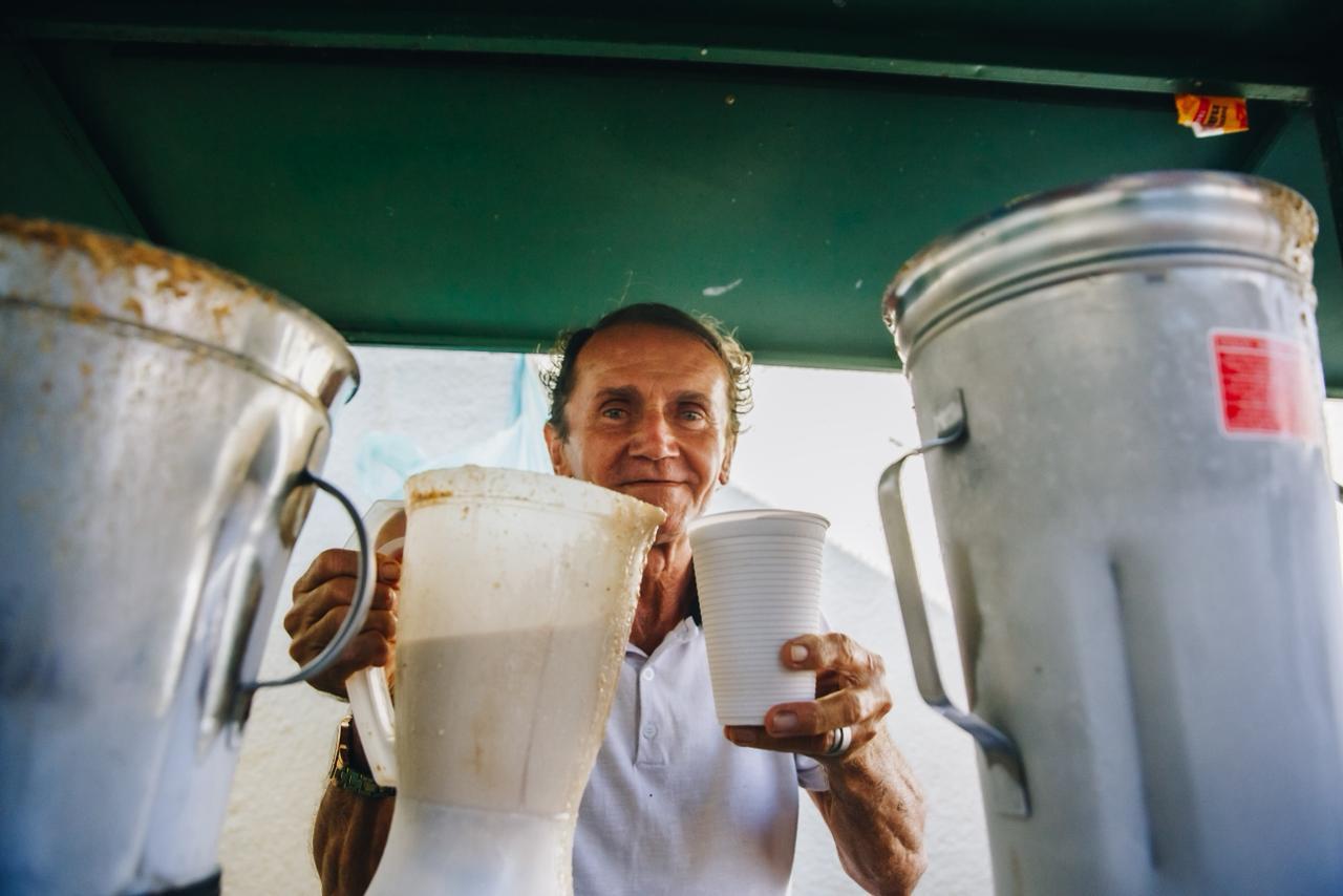 Na imagem, uma foto de ângulo baixo mostra um homem de pele morena clara, com cabelos escuros e encaracolados e um sorriso, segurando um copo descartável branco e um liquidificador de plástico com uma bebida cor de bege ou marrom claro, tipo milk-shake. Ele está em um quiosque ou barraca, com uma cobertura verde acima dele, e está usando uma camisa polo branca. O homem estende o copo na direção da câmera. À esquerda e à direita do homem, parcialmente visíveis em primeiro plano, estão grandes liquidificadores industriais de metal prateado, prontos para uso. A bebida é provavelmente guaraná batido, um popular drink brasileiro.