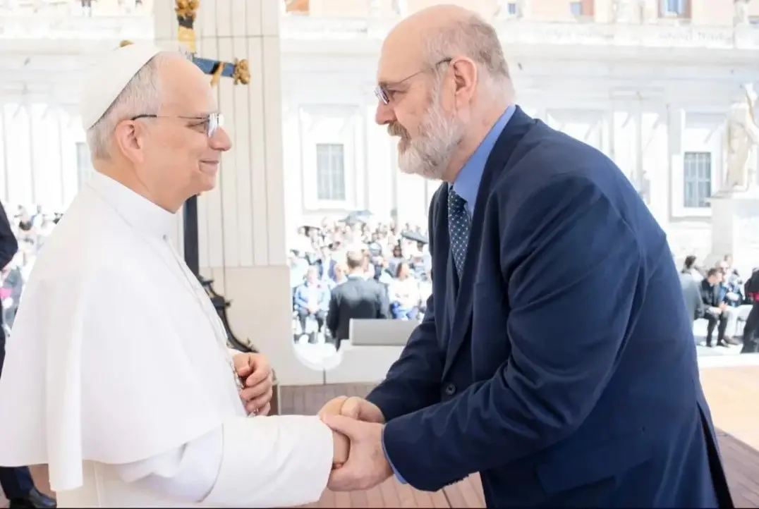 Silvonei José cumprimenta o Papa Leão XIV em uma cerimônia ao ar livre no Vaticano. Ambos sorriem e trocam um aperto de mãos cordial. O locutor brasileiro veste terno azul e gravata, enquanto o Papa está de branco, usando solidéu. Ao fundo, fiéis e convidados acompanham o encontro na Praça de São Pedro.