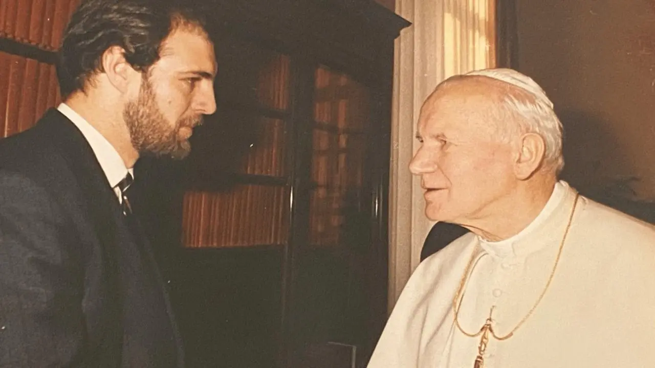 Em uma imagem de tom amarelado, Silvonei José, ainda jovem, encontra o Papa João Paulo II. Os dois estão em perfil, trocando olhares atentos e próximos, num ambiente interno do Vaticano. O Papa veste batina branca e crucifixo dourado; Silvonei usa terno escuro e gravata.
