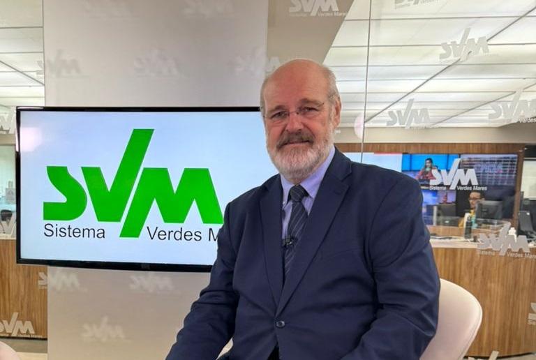 Silvonei José posa sorridente em um estúdio moderno do Sistema Verdes Mares, em Fortaleza. Atrás dele, uma tela exibe o logotipo “SVM” em verde. Ele está de terno azul e gravata, com microfone preso ao paletó, transmitindo serenidade e profissionalismo. O ambiente é iluminado e reflete o clima de redação jornalística.