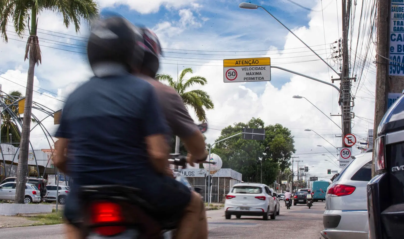 Motociclista borrado em primeiro plano em uma rua urbana. O foco está em um sinal de trânsito elevado no centro que indica ATENÇÃO VELOCIDADE MÁXIMA PERMITIDA: 50 km/h.