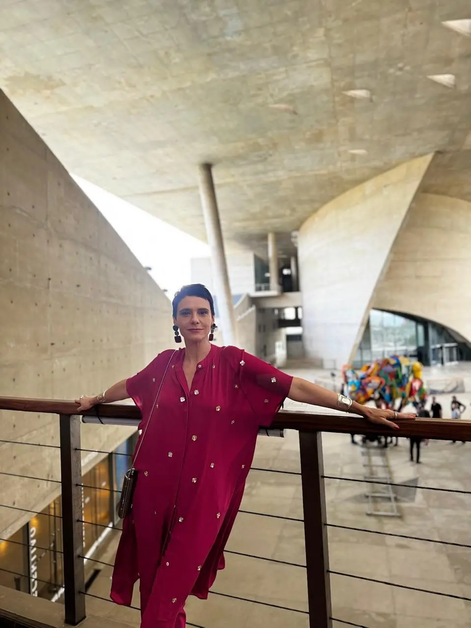 Imagem mostra uma mulher, a atriz Malu Galli, com os braços abertos e as mãos apoiadas em um guarda corpo. Ela está vestindo uma roupa vermelha e usa uma pequena bolsa preta de lado.