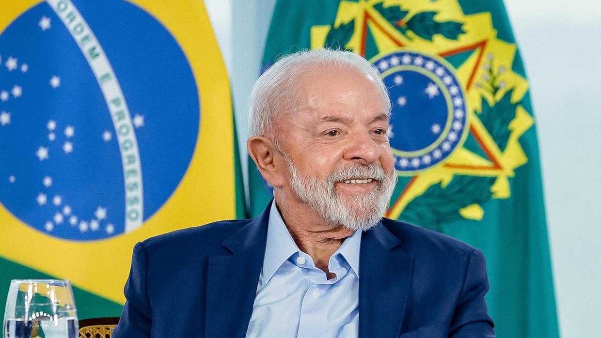 Lula sorri durante evento oficial, com bandeiras do Brasil ao fundo.