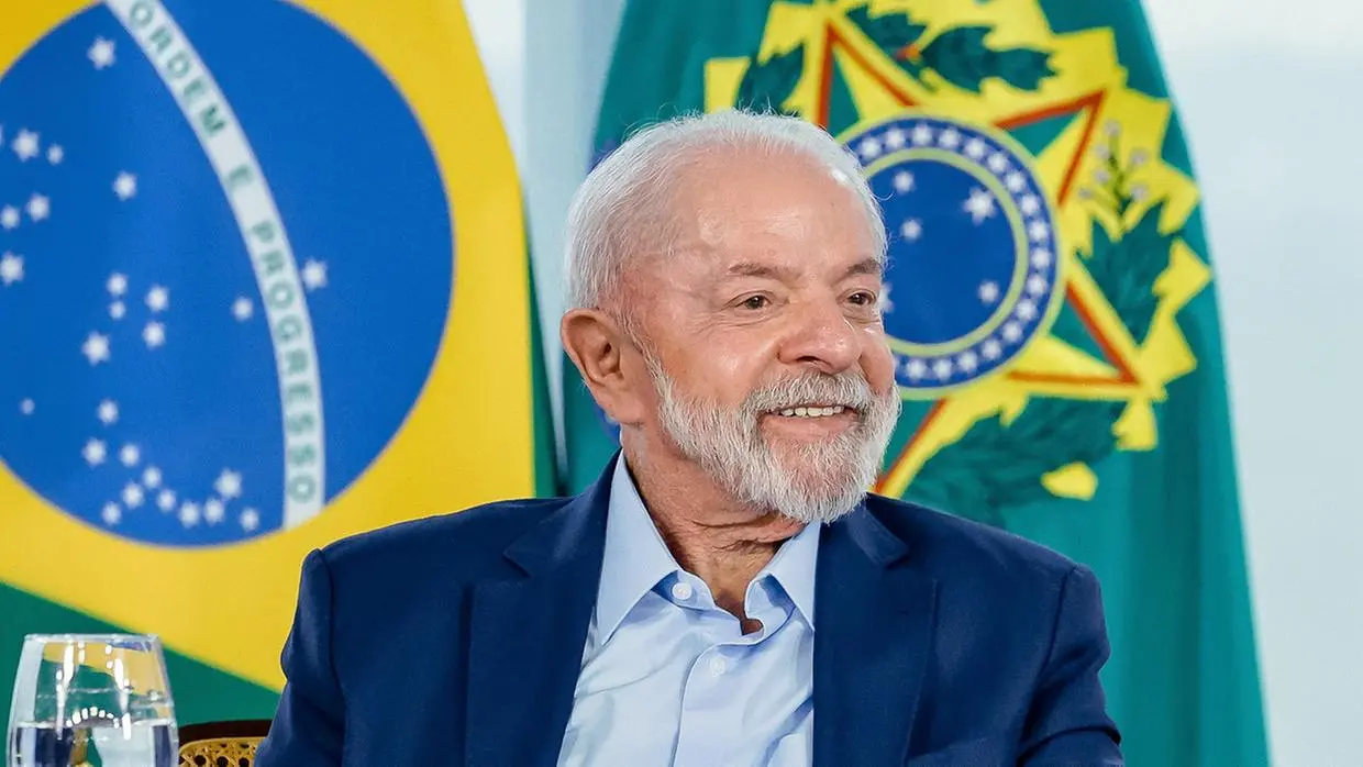 Lula sorri durante evento oficial, com bandeiras do Brasil ao fundo.