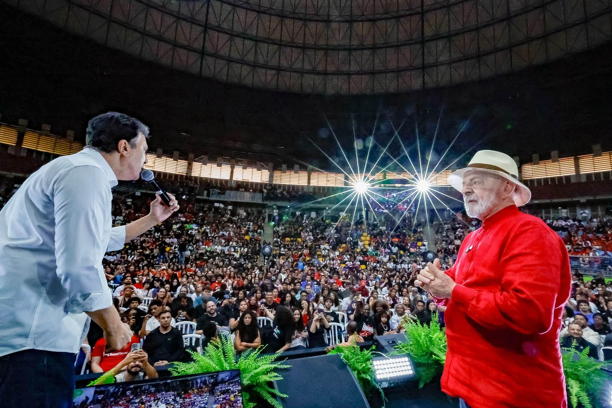 Lula observa Camilo discursando. Ambos estão em cima do palco. Camilo veste branco, Lula, vermelho. Ao mundo, milhares de estudantes acompanham o evento