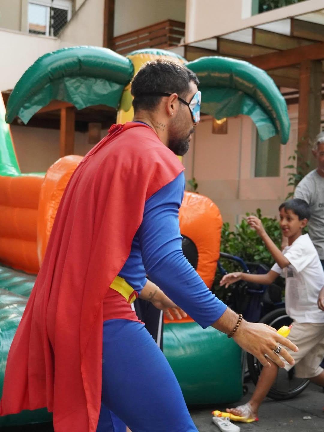 Imagem mostra homem de costas. Ele está fantasiado de Superman, está vendado e aparentemente brincando com crianças. Uma criança aparece à direita e há um brinquedo inflável ao fundo.