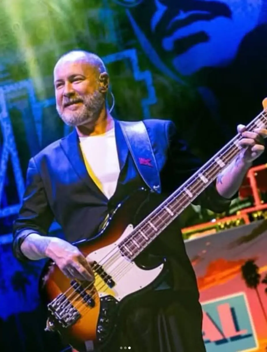 Baixista Sam Rivers, do Limp Bizkit, no palco. Ele é um homem de pele clara, barba grisalha e cabeça raspada, vestindo um blazer preto com forro amarelo e uma camiseta branca, enquanto toca um baixo elétrico sunburst de quatro cordas.