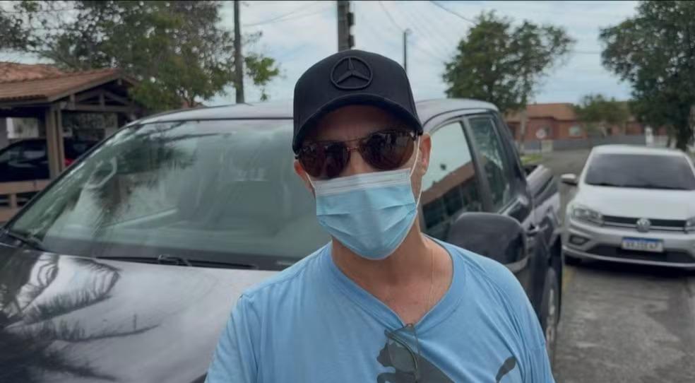 Arnaldo Augusto é um homem branco e idoso. Na foto, ele usa boné preto, óculos escuros, máscara cirúrgica azul e uma camiseta também azul.