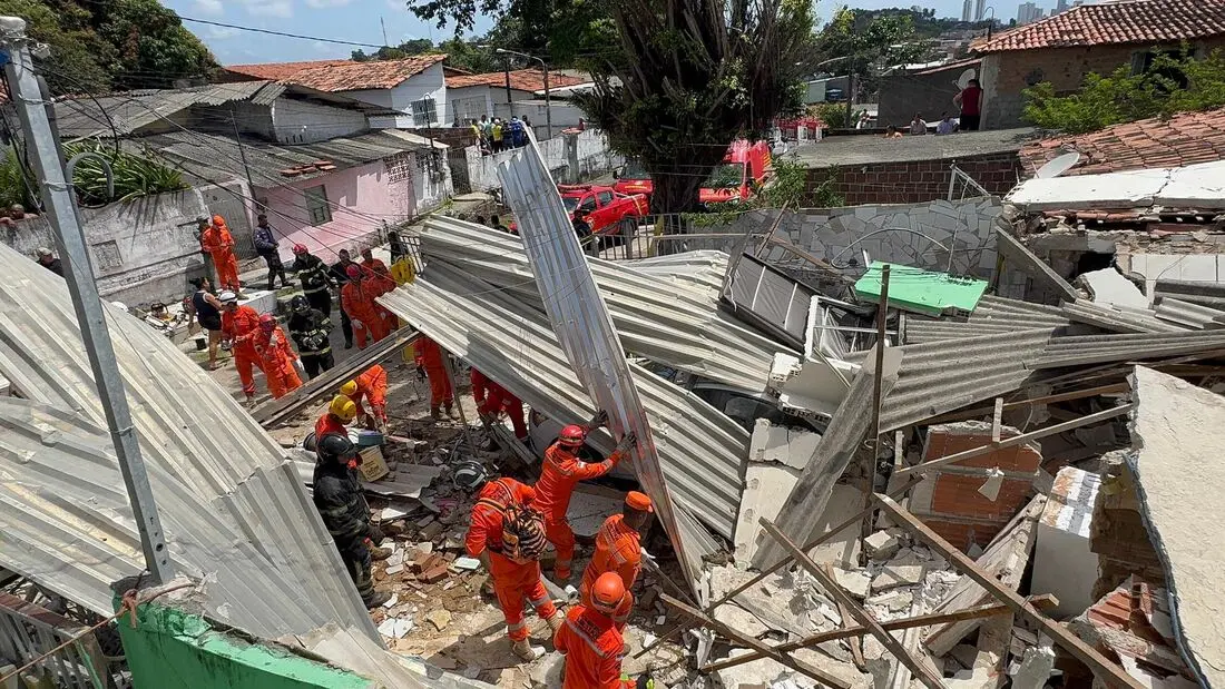Equipes do Corpo de Bombeiros, usando uniformes laranja e capacetes, trabalham entre os escombros de casas destruídas após uma explosão em Olinda (PE). Alguns bombeiros removem placas metálicas e destroços, enquanto outros observam e prestam apoio na área. O local está tomado por entulho, pedaços de telhado e paredes desabadas. Ao fundo, há outras residências e veículos de emergência estacionados na rua, sob um céu claro.
