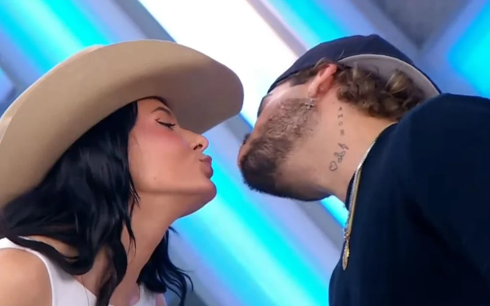 Dois artistas trocam um selinho durante uma gravação de programa de TV. A mulher, de cabelos escuros e chapéu de cowboy bege, se inclina para frente com os lábios estendidos. O homem, usando boné virado para trás e colar dourado, também se aproxima para o beijo. O fundo exibe luzes e painéis azulados típicos de um estúdio televisivo.