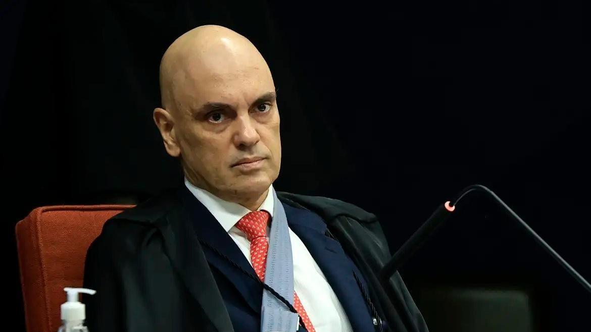 Alexandre de Moraes é um homem branco, de meia idade e careca. Na foto, ele está de terno preto, camisa social branca e gravata vermelha. Seu semblante é sério.