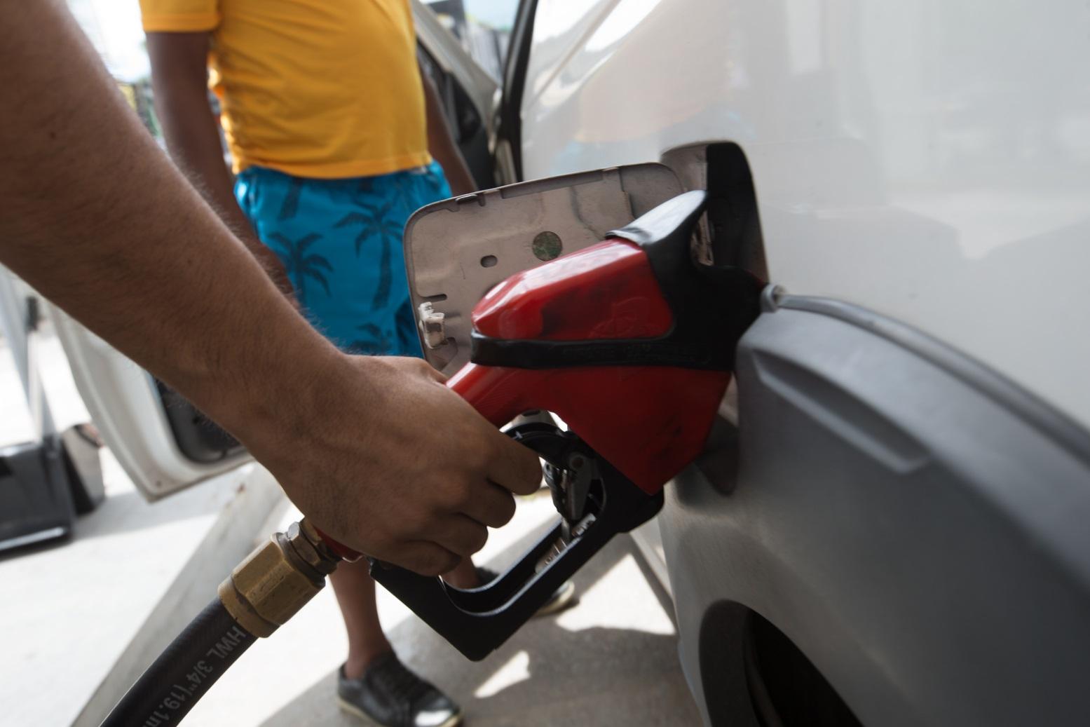 Um homem segura o bico de uma bomba de combustível vermelha e abastece um carro branco em um posto de gasolina. Outro homem, de camiseta amarela e bermuda azul, aparece ao fundo.