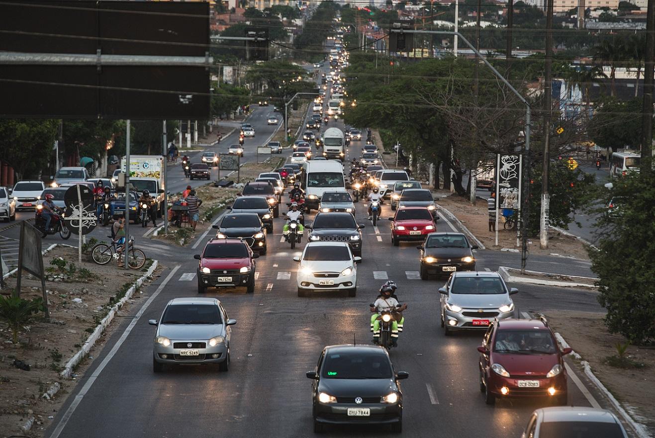 Avenida Mister Hull, em Fortaleza, movimentada com carros e motos em várias faixas, com o trânsito intenso e faróis acesos num fim de tarde. Pessoas circulam nas calçadas e canteiros laterais, e há árvores e prédios ao fundo.