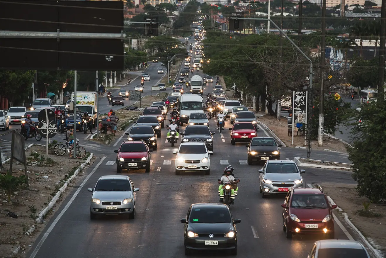Avenida Mister Hull, em Fortaleza, movimentada com carros e motos em várias faixas, com o trânsito intenso e faróis acesos num fim de tarde. Pessoas circulam nas calçadas e canteiros laterais, e há árvores e prédios ao fundo.