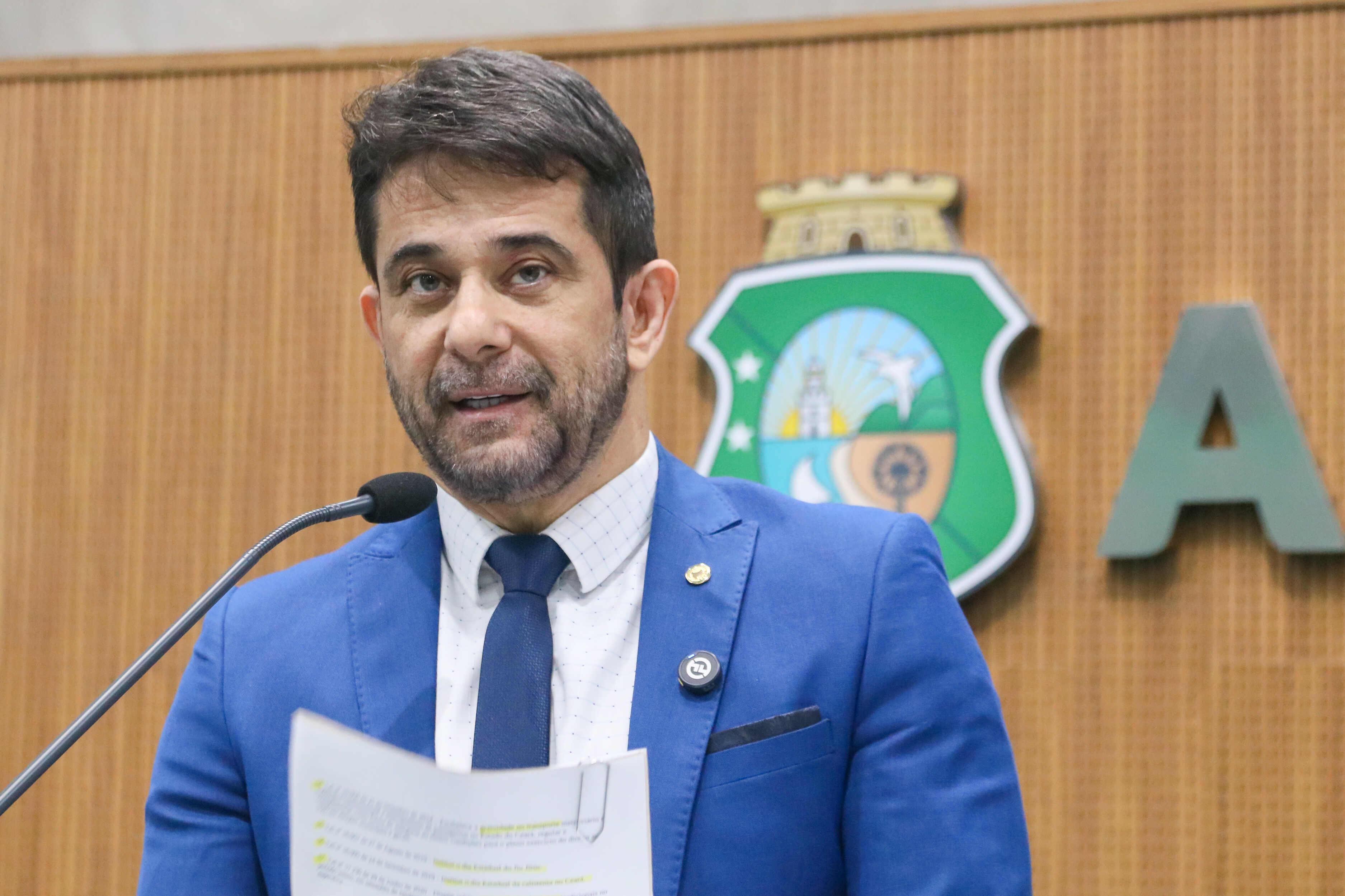 Homem de terno azul, camisa branca e gravata azul escura fala ao microfone em um plenário. Ele segura um papel com anotações impressas. Ao fundo, há parte de um brasão nas cores verde, azul e amarelo e letras em relevo na parede de madeira.