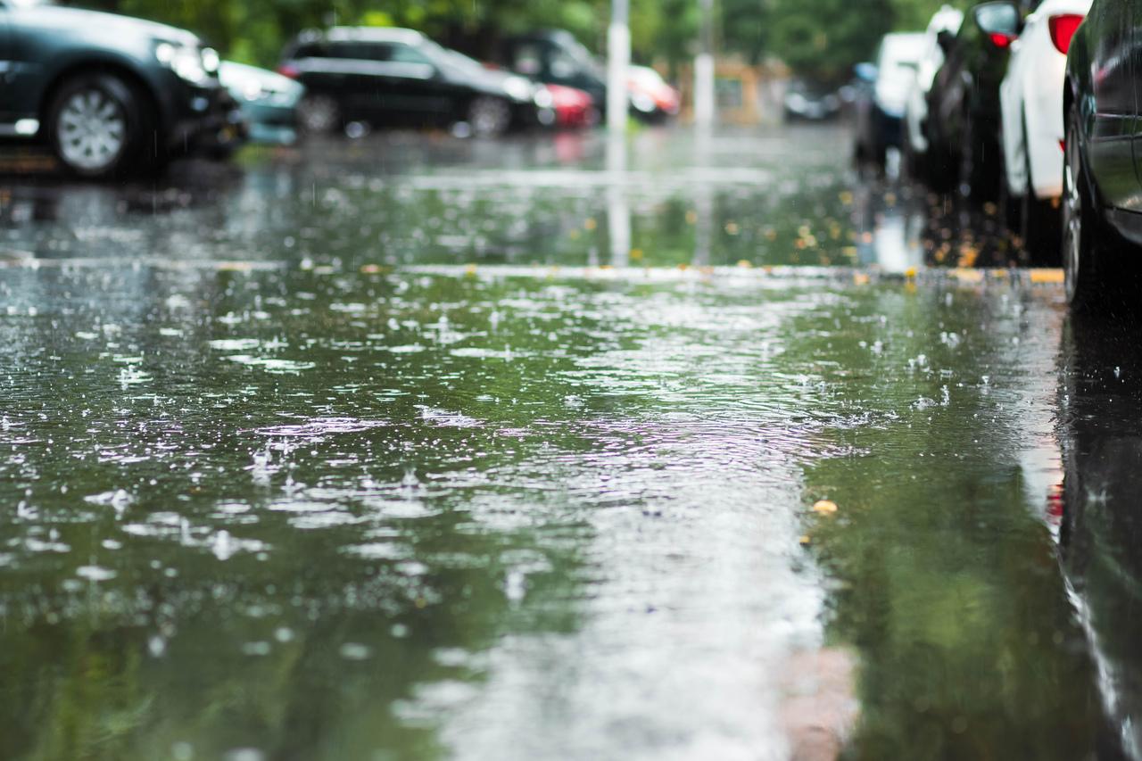 Estacionamento alagado devido à forte chuva, com carros parcialmente submersos em uma rua molhada e reflexos na água.