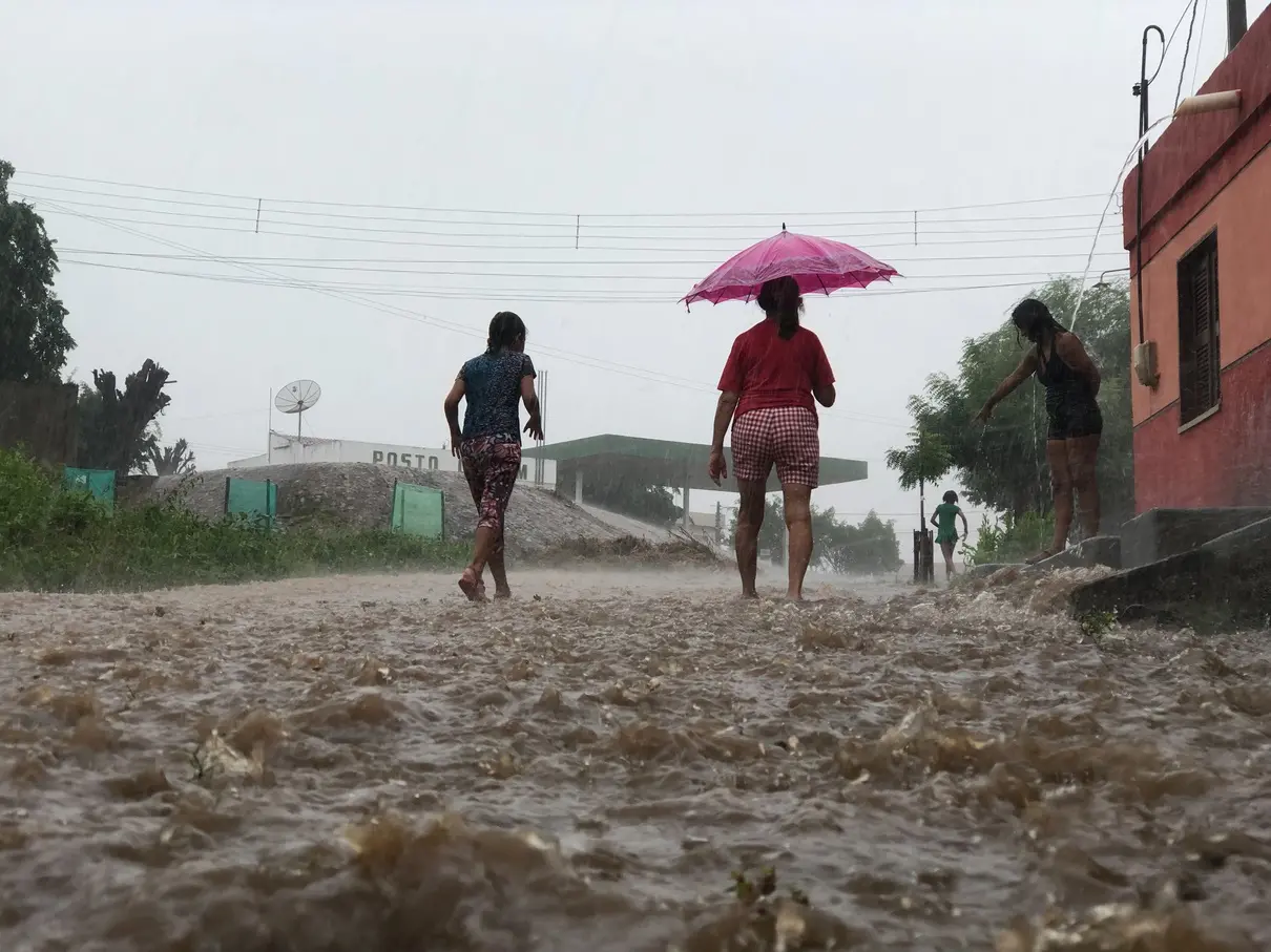 Em uma rua de terra alagada pela chuva forte em Iguatu, no interior do Ceará, três mulheres caminham com os pés na água barrenta; uma delas segura um guarda-chuva rosa. À direita, uma mulher observa da calçada enquanto a enxurrada desce pela via. Ao fundo, há fios de energia e algumas árvores.