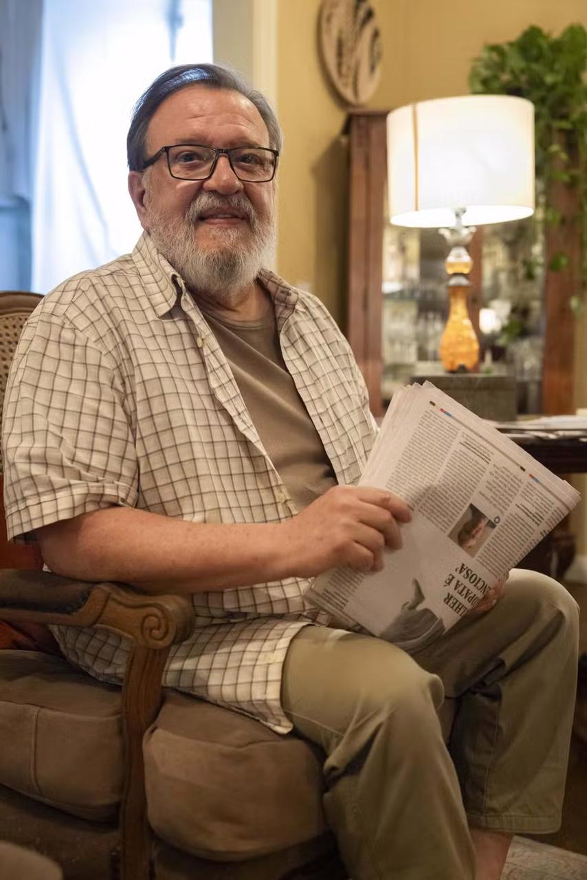 Ator Luis Melo, caracterizado de Bartolomeu para remake de Vale Tudo. Sorridente com barba, óculos, usando camiseta xadrez e lendo jornal em sala de estar aconchegante com iluminação suave e decoração moderna.