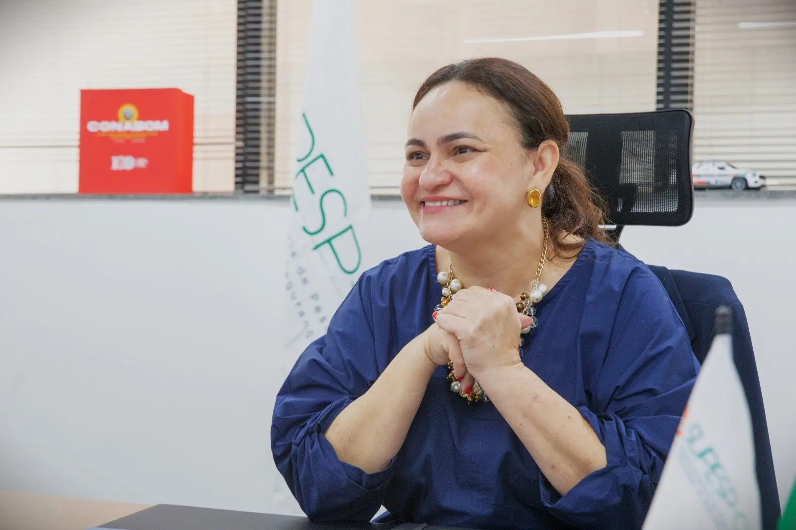 Juliana Barroso, superintendente da Supesp no Ceará, sorridente em ambiente de escritório, usando blusa azul, com acessórios de joias e sentada em cadeira de escritório, praticando postura confiante.