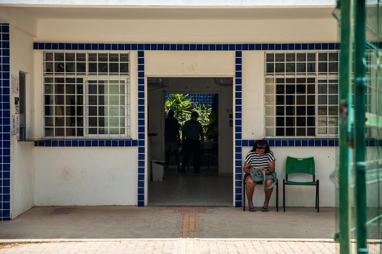 Fachada branca com detalhes em azul mostra a entrada de uma unidade de saúde. Uma mulher senta em uma cadeira verde na calçada, enquanto duas pessoas são vistas no interior do prédio.