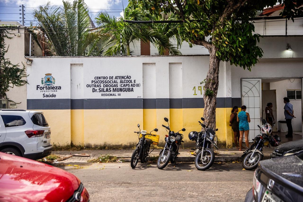 Fachada amarela e cinza da unidade Dr. Silas Munguba, com motos estacionadas em frente e pessoas conversando na entrada. O logotipo da Prefeitura de Fortaleza aparece pintado na parede.
