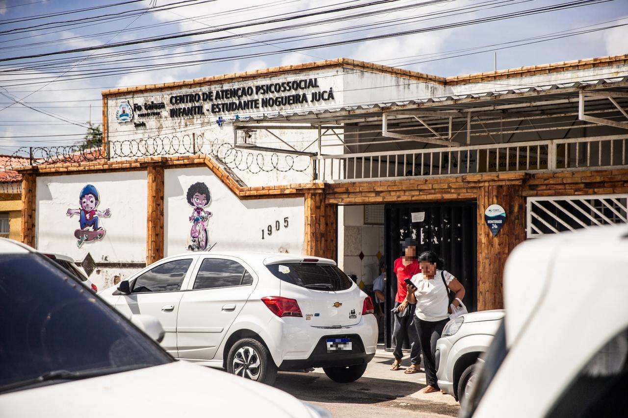 Entrada de Caps Infantil com muro branco e detalhes em tijolos aparentes, decorado com desenhos infantis coloridos. Carros estacionados em frente e pessoas entrando e saindo do local.