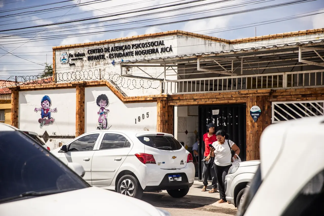 Entrada de Caps Infantil com muro branco e detalhes em tijolos aparentes, decorado com desenhos infantis coloridos. Carros estacionados em frente e pessoas entrando e saindo do local.